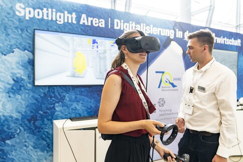 A young woman operates a presentation with VR glasses at a trade fair stand, her progress is visible on a screen in the background.