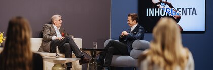 Two men in suits are sitting in armchairs on a stage at INHORGENTA, chatting in front of an audience.