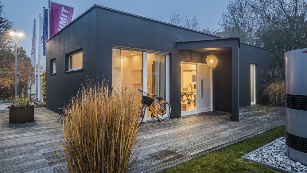 Modernes würfelförmiges Gebäude mit großen Fenstern, umgeben von Gras und Pflanzen, unter einem bewölkten Abendhimmel.