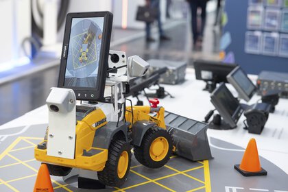 A yellow model bulldozer with a screen stands on a table, surrounded by orange-coloured hats and other equipment.