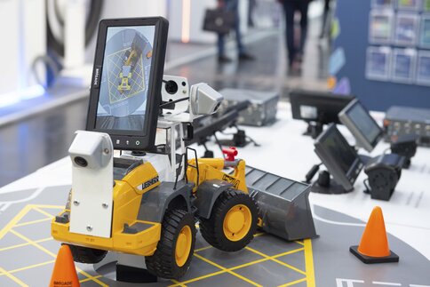 A yellow model bulldozer with a screen stands on a table, surrounded by orange-coloured hats and other equipment.