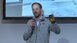 Person in grey jumper gives a lecture with microphone in front of a presentation.