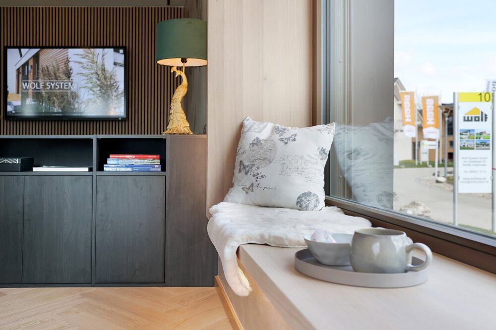 Eine moderne Fensterbank mit Kissen und Decke zum Sitzen. Im Hintergrund hängt ein Fernseher und davor steht ein schwarzes Regal mit einer vergoldeten Lampe darauf.