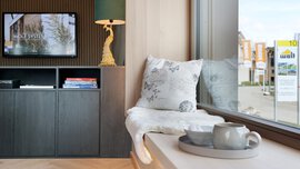 Eine moderne Fensterbank mit Kissen und Decke zum Sitzen. Im Hintergrund hängt ein Fernseher und davor steht ein schwarzes Regal mit einer vergoldeten Lampe darauf.