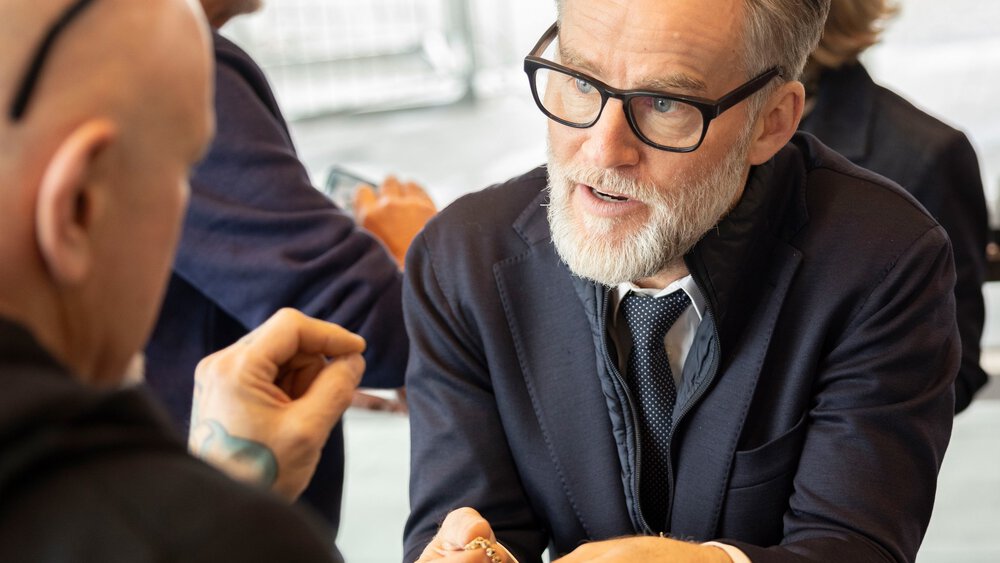 A bearded man with glasses discusses a piece of gold jewellery at a table with another person in a suit.