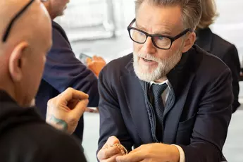 Ein bärtiger Mann mit Brille bespricht an einem Tisch mit einer anderen Person im Anzug ein goldenes Schmuckstück.