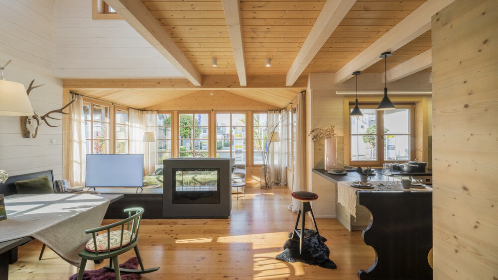 Gemütliches Wohnzimmer mit Holzbalken, Kamin und großen Fenstern, die Tageslicht hereinlassen.