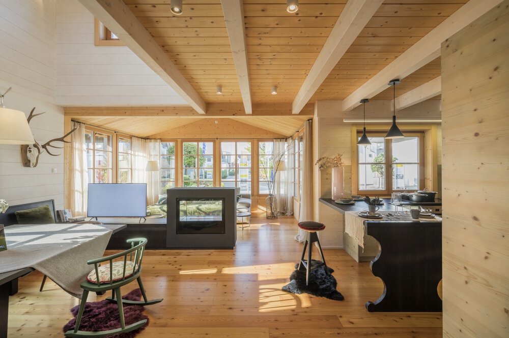 Gemütliches Wohnzimmer mit Holzbalken, Kamin und großen Fenstern, die Tageslicht hereinlassen.