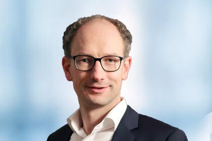 Portrait of a man with glasses, short hair, and dark suit against a blue-white background.