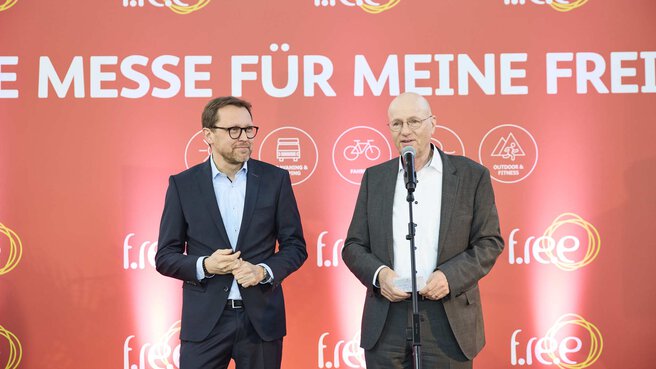 Zwei Männer stehen bei einer Eröffnungsfeier auf einer Bühne vor einer Leinwand. Ein Mann spricht in ein Mikrofon.