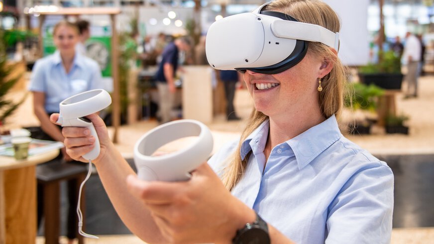A smiling woman wearing a white VR headset and holding up the controllers at a trade fair stand.