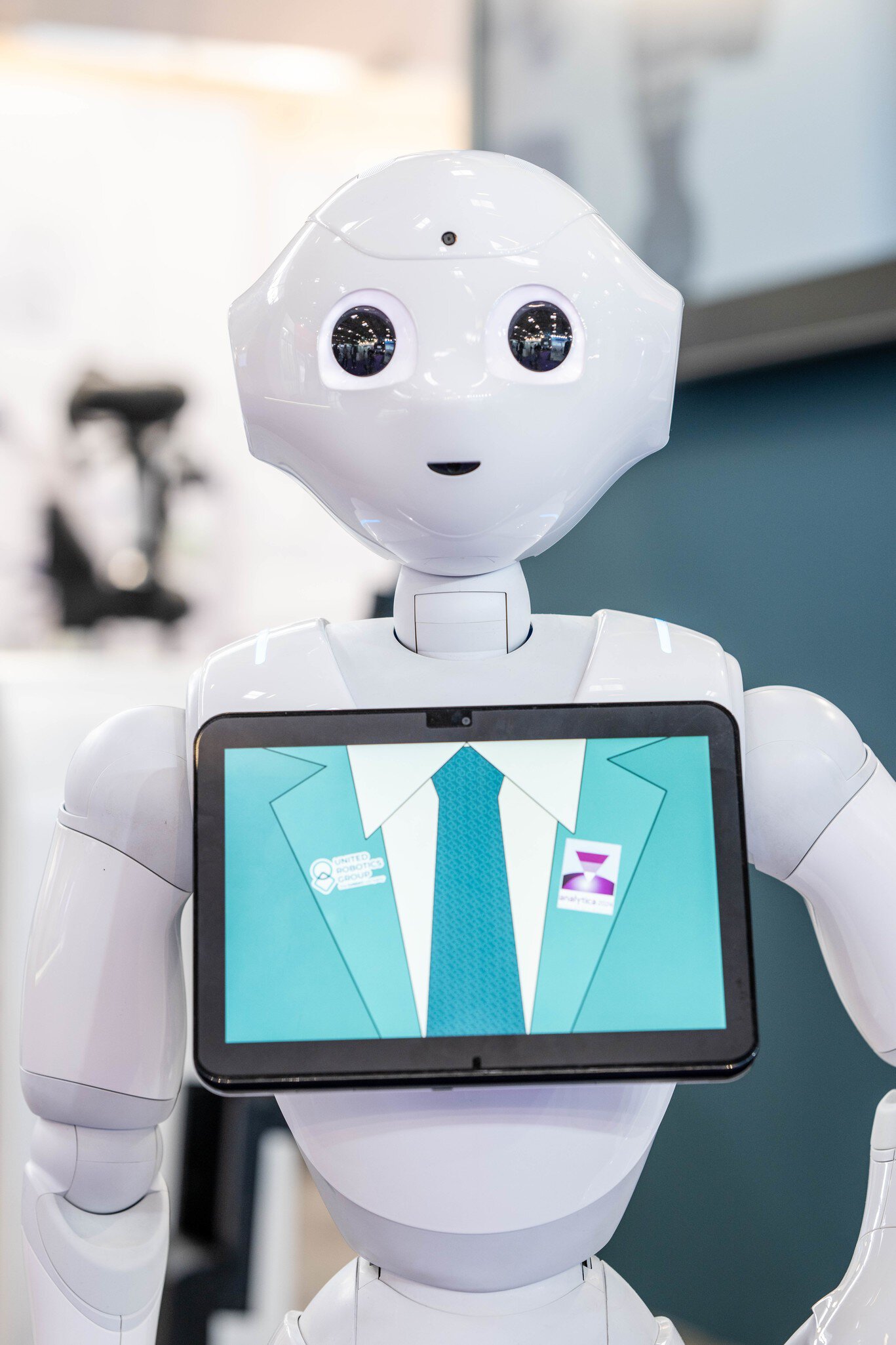 A humanoid robot with a white plastic casing has a screen on its chest. The screen shows an illustration of a suit and tie and the analytica logo.