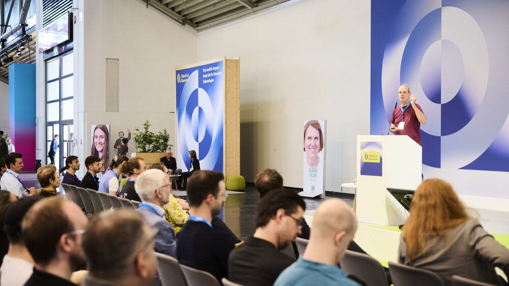 Ein Redner spricht auf der World of Quantum in München vor Publikum, umgeben von Bannern und Porträts zur Quantenforschung.