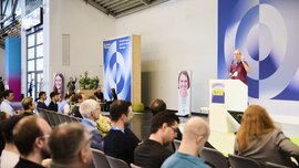 Ein Redner spricht auf der World of Quantum in München vor Publikum, umgeben von Bannern und Porträts zur Quantenforschung.