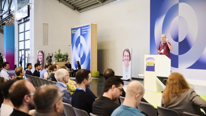 Ein Redner spricht auf der World of Quantum in München vor Publikum, umgeben von Bannern und Porträts zur Quantenforschung.
