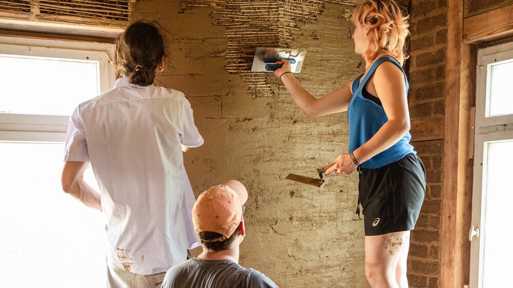 Drei Personen auf Leitern beim Verputzen einer Wand in einem zu renovierenden Raum, mit Werkzeugen und Eimern in der Nähe.