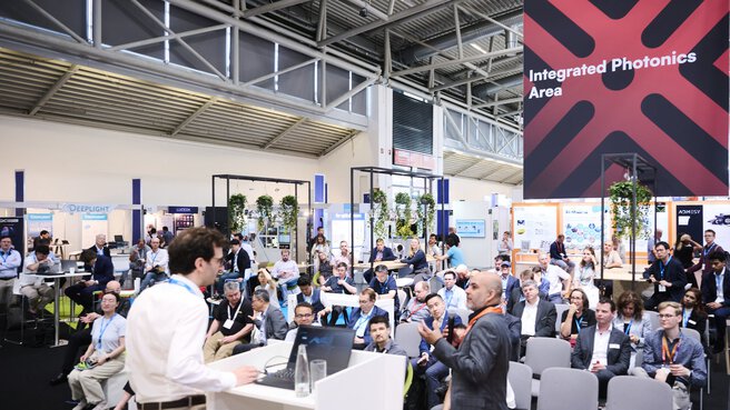 Presentation at the Laser World of Photonics trade show in front of an audience, with exhibition booths and a sign reading “Integrated Photonics Area” in the background