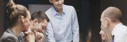 A work team in a work meeting. A woman in a shirt blouse is presenting something to her four colleagues