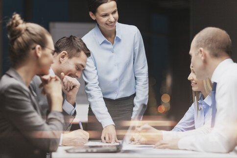 A work team in a work meeting. A woman in a shirt blouse is presenting something to her four colleagues