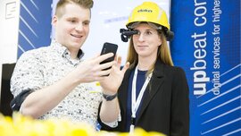 Zwei Aussteller stehen an einem Stand auf der digitalBAU. Er hält ein Smartphone in der Hand, sie trägt einen Bauarbeiterhelm mit einer Vorrichtung vor dem einen Auge.