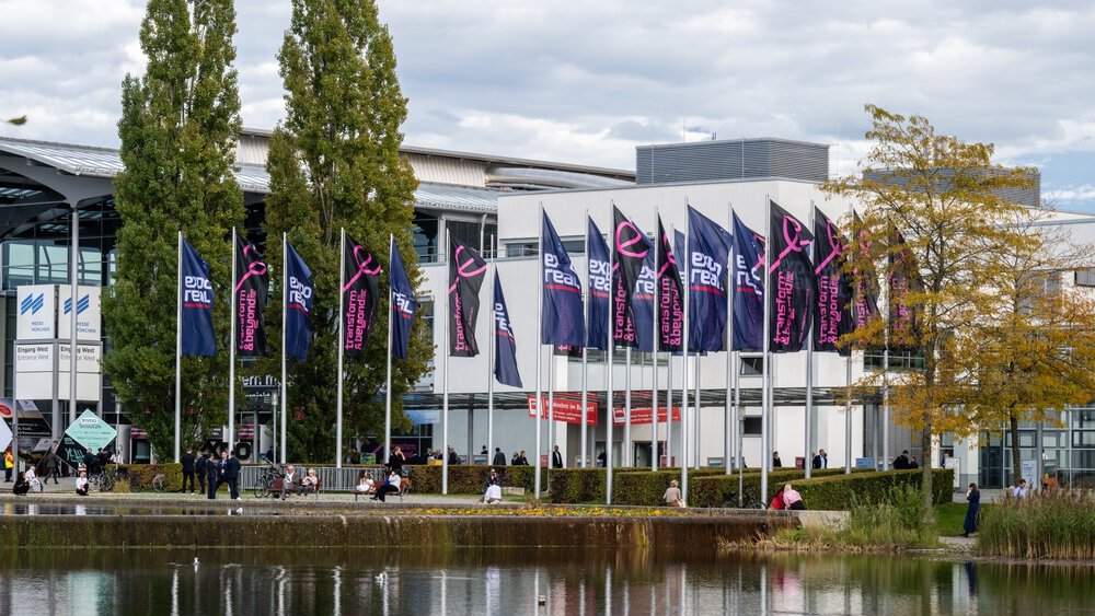 Zahlreiche Fahnenmasten mit Fahnen der EXPO REAL stehen vor dem Eingang West des Messe München Geländes. Im Vordergrund ist der Messesee zu sehen.