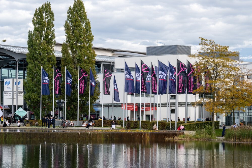 Numerous flagpoles with EXPO REAL flags stand in front of the West Entrance to the Messe München exhibition centre. The trade fair lake can be seen in the foreground.