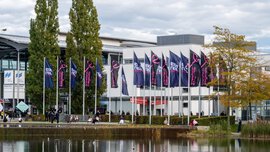 Numerous flagpoles with EXPO REAL flags stand in front of the West Entrance to the Messe München exhibition centre. The trade fair lake can be seen in the foreground.