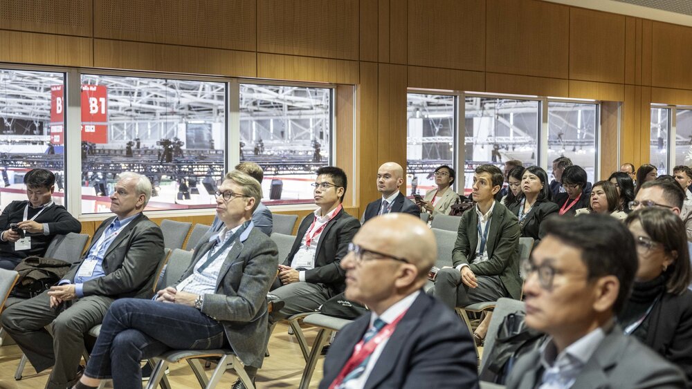 Teilnehmerinnen und Teilnehmer einer Fachkonferenz auf der EXPO REAL in München sitzen in einem Seminarraum und hören aufmerksam einem Vortrag zu. Durch die großen Fenster im Hintergrund ist das Messegelände mit der Beschilderung der Hallen zu sehen. Die Atmosphäre ist konzentriert und professionell.
