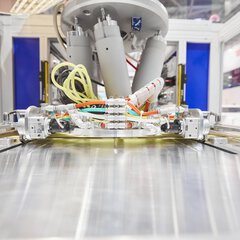 Kleine Industriemaschine mit grauen Zylindern und bunten Kabeln wird während der productronica in einem Schaukasten präsentiert.