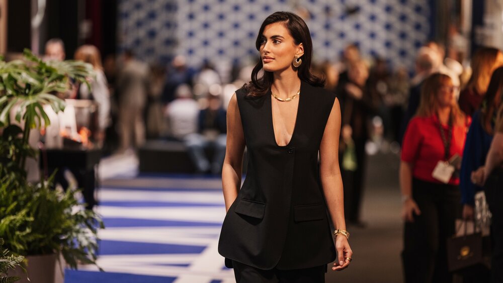 A smiling woman wearing a low-cut black suit and gold jewelry on her arms, neck, and ears walks past the exhibition booths.