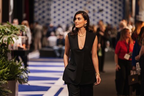 A smiling woman wearing a low-cut black suit and gold jewelry on her arms, neck, and ears walks past the exhibition booths.