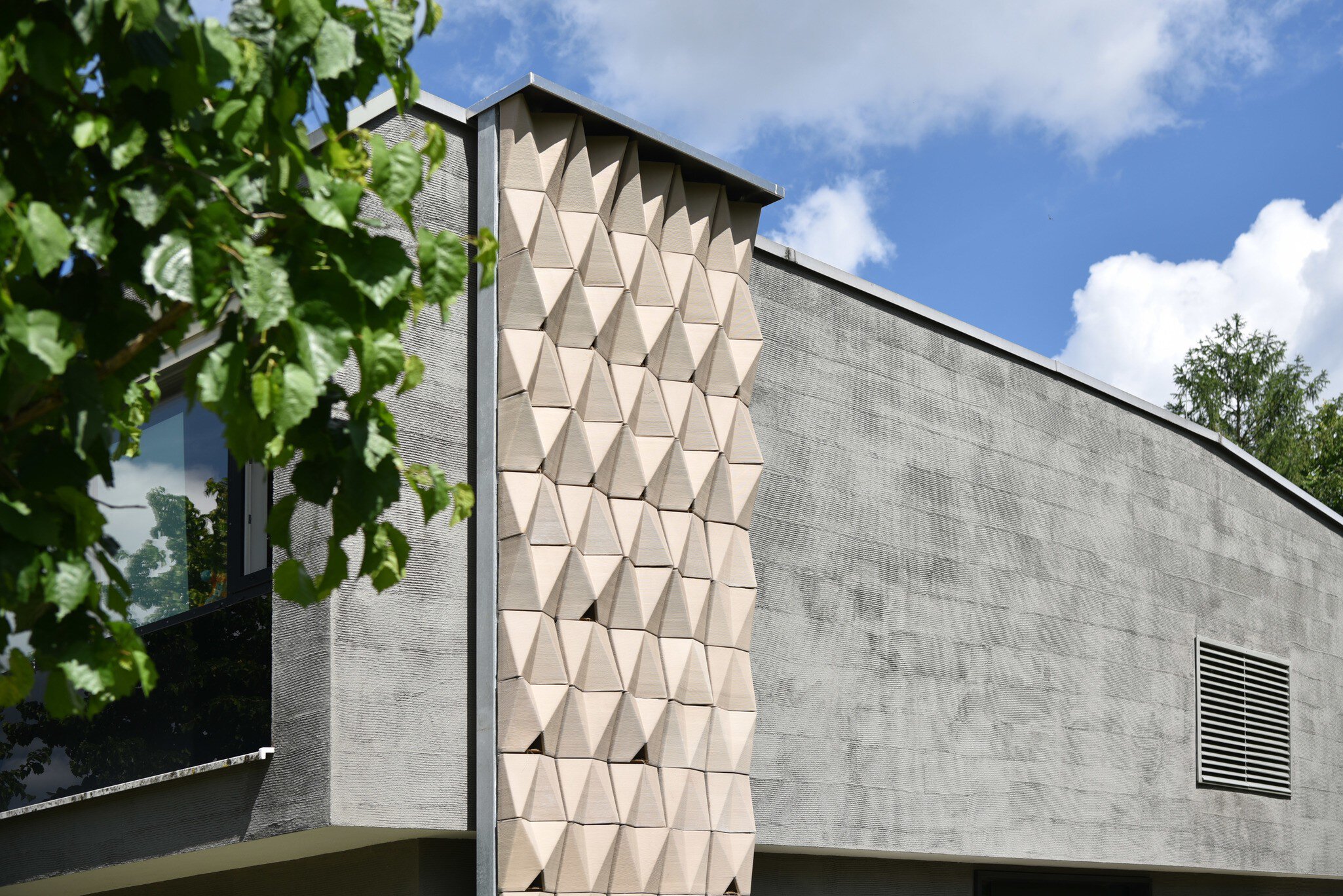 Modern gray building with geometrically structured panels and tree branches in the foreground under a blue sky.