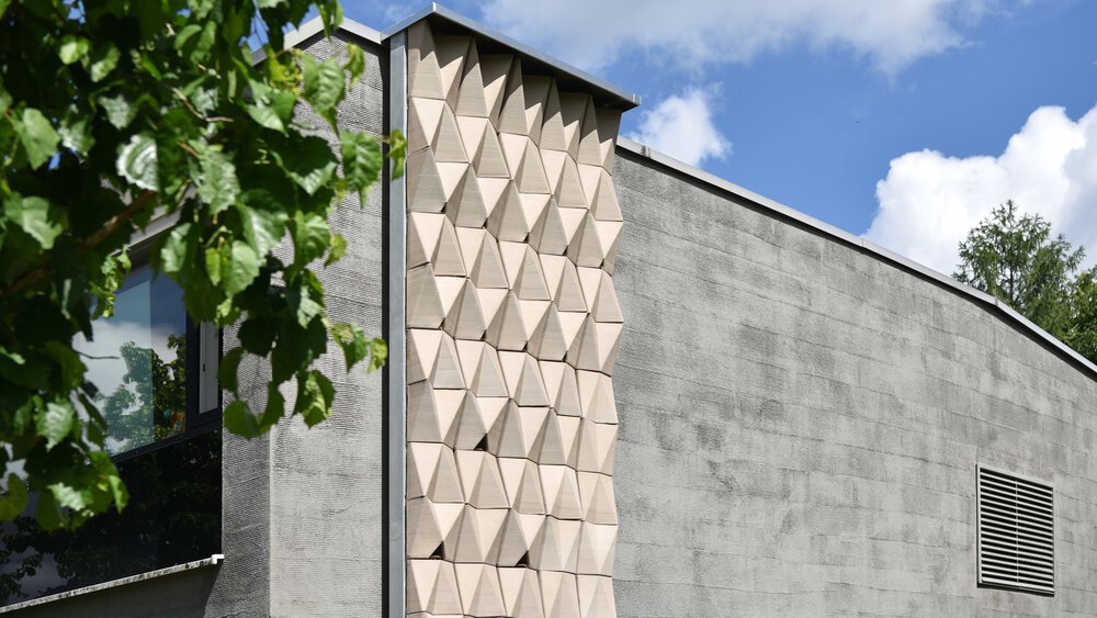 Modern gray building with geometrically structured panels and tree branches in the foreground under a blue sky.