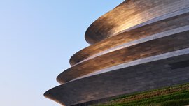 A futuristic building consisting of several levels sits atop a terraced green slope and reflects silver in the evening sun.