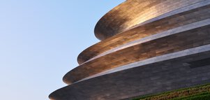 A futuristic building consisting of several levels sits atop a terraced green slope and reflects silver in the evening sun.