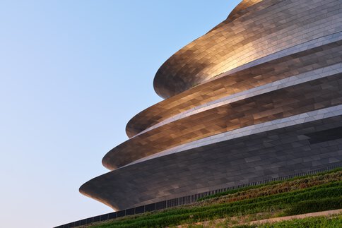 Ein futuristisches Gebäude, bestehend aus mehreren Ebenen, liegt über einem stufigen begrünten Hang und reflektiert silbern in der Abendsonne.