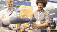 Two people at a trade fair stand are interested in a robot arm and smiling.