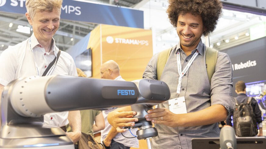 Two people at a trade fair stand are interested in a robot arm and smiling.