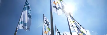 Flaggen mit den Logos der Messe München und der IFAT Munich wehen an Masten vor einem sonnigen blauen Himmel.
