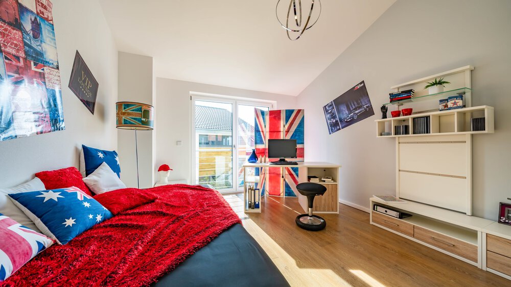 Ein helles Schlafzimmer mit einer roten Tagesdecke, Union-Jack-Dekor, einem Schreibtisch mit Computer, Bücherregalen und großen Fenstern, durch die natürliches Sonnenlicht hereinströmt.