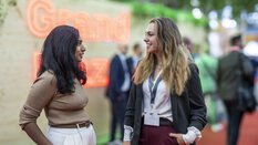 Two women are having a friendly conversation at EXPO REAL in Munich. Both are dressed in business attire and smiling at each other. In the blurred background, a glowing “Grand Plaza” sign and other trade fair visitors can be seen. The scene conveys an open and professional networking atmosphere.