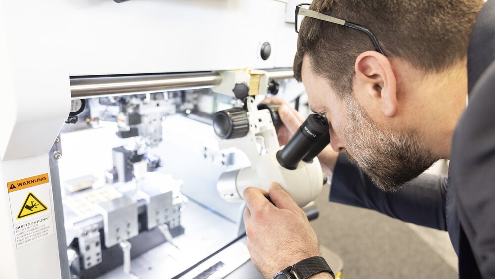 Ein Mann mit braunen Haaren und Brille auf dem Kopf blickt durch ein Mikroskop, welches auf das Innere einer weißen geöffneten Maschine gerichtet ist.