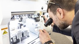Ein Mann mit braunen Haaren und Brille auf dem Kopf blickt durch ein Mikroskop, welches auf das Innere einer weißen geöffneten Maschine gerichtet ist.
