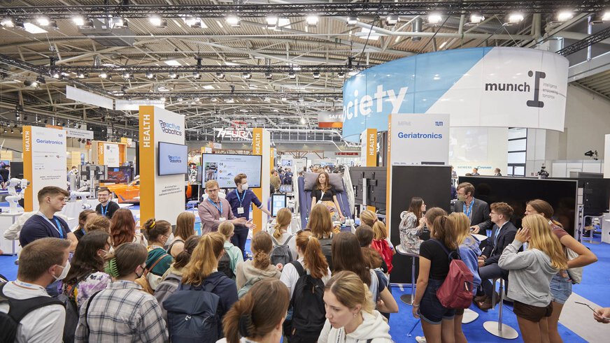 Eine Gruppe junger Menschen nimmt an einer Demonstration an einem Messestand teil. Im Hintergrund sind weitere Stände und Menschen zu sehen.