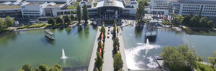 Luftaufnahme des Messesees und dem Eingang West. Ein breiter Weg führt, gesäumt von Bäumen, Flaggen und Wasser zur Eingangshalle.