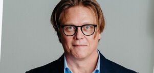 Picture of a man with brown, half-length hair and black glasses, wearing a dark blue jacket with a light blue shirt.