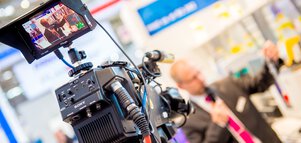 A camera from the perspective of a cameraman. A man with a microphone stands in front of the camera and presents something standing in an exhibition hall.