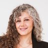 A smiling woman with curly hair that is half grey and half brown is wearing earrings, a necklace, a light blue top and a black suit.
