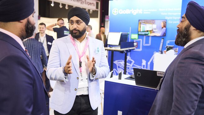 Drei Männer in Anzügen führen auf der EXPO REAL ein Gespräch am Messestand von „GoBright“. Der Mann in der Mitte gestikuliert beim Erklären, während die anderen aufmerksam zuhören. Im Hintergrund sind Bildschirme mit digitalen Arbeitsplatzlösungen und Messeteilnehmende zu sehen.
