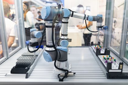 An automated, blue-grey robotic arm in the laboratory environment at the analytica trade fair. It is positioned in a workstation equipped with sample holders. Some people can be seen out of focus in the background.